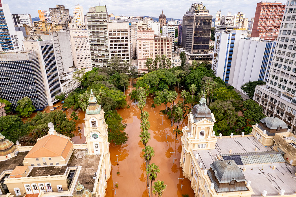 Cidades do Futuro Como preparar as cidades para os desafios do futuro na gestão de desastres e emergências Márcia Casseb, engenheira civil, especialista em desenvolvimento territorial e urbano e mestre em saneamento, meio ambiente e recursos hídricos. A pandemia de COVID-19 foi declarada pela Organização Mundial da Saúde (OMS) em março de 2020, tornando necessário o distanciamento social e o isolamento para evitar a propagação da doença e possibilitar o retorno à normalidade. Na América Latina e no Caribe (ALC), os meses de confinamento duraram, em média, entre cinco e sete meses. Esse período trouxe inúmeros desafios em diversos setores, afetando de forma distinta a vida das pessoas. Naquela ocasião, eu trabalhava no Banco Interamericano de Desenvolvimento (BID), no Paraguai, atuando principalmente em projetos urbanos em áreas informais. As famílias que viviam nessas comunidades enfrentaram grandes dificuldades para se adaptar à nova realidade o isolamente em casas pequenas e superlotadas, a impossibilidade de realizar trabalho remoto, as limitações de conectividade para dar continuidade às atividades profissionais e aos estudos de crianças, adolescentes e adultos — muitas vezes com apenas um computador disponível ou sem acesso a esse equipamento. Os problemas foram múltiplos e complexos, e governos, junto a diferentes instituições, mobilizaram-se para apoiar as comunidades mais vulneráveis que necessitavam de auxílio. Uma pandemia é uma emergência de saúde pública, mas outros tipos de desastres podem ocorrer a qualquer momento. Chuvas intensas podem provocar inundações, enchentes e deslizamentos, afetando principalmente populações vulneráveis que vivem em áreas de risco, Cidades localizadas em regiões áridas ou semiáridas podem enfrentar períodos prolongados de seca sem contar com instrumentos de segurança hídrica, ou sofrer com temperaturas extremas que geram ondas de calor, aumentando o consumo de água e energia e impactando a saúde — sobretudo de idosos e crianças. Diante desse cenário, diversas instituições nacionais e internacionais, universidades e especialistas discutiram e apoiaram medidas para fortalecer as cidades, de modo que estejam mais preparadas e resilientes diante desses eventos. A Divisão de Habitação e Desenvolvimento Urbano (HUD), com o apoio da Divisão de Meio Ambiente, Desenvolvimento Rural e Gestão de Desastres (ENR) do BID, mobilizou-se para oferecer assistência técnica a governos locais dos países do Cone Sul (Argentina, Chile, Paraguai e Uruguai), incentivando-os a pensar estrategicamente sobre o futuro. A ideia central do trabalho foi impulsionar estratégias para que cada cidade se conheça, saiba gerir seus riscos, compreenda os processos que enfrenta, desenvolva planos de emergência e promova um futuro resiliente. Do trabalho extenso de orientação técnica participativa realizado com 16 municípios da região, foi possível adaptar um índice previamente desenvolvido pelo Banco, voltado a medir a qualidade das políticas públicas de gestão de riscos de desastres, desta vez com enfoque no nível local o Índice de Governança Local e Políticas Públicas para Gestão de Riscos (iLGOPP Local). Esse índice é uma ferramenta concebida para apoiar a avaliação de um conjunto de condições regulatórias, institucionais e orçamentárias consideradas fundamentais para a implementação de processos de gestão de riscos de catástrofes em âmbito local. Dessa experiência resultou a publicação em espanhol “Ciudades a Prueba de Futuro”” que define a gestão de riscos da seguinte forma (em tradução livre) “O que é GRD A gestão de riscos de desastres (GRD) refere-se a todos os processos destinados a conceber, implementar e avaliar estratégias, políticas e medidas que visem melhorar a compreensão dos riscos de desastres; promover a redução, mitigação e transferência desses riscos; e promover a melhoria contínua das práticas de preparação, resposta, recuperação e adaptação às mudanças climáticas. Seu objetivo explícito é aumentar a segurança humana, o bem-estar, a qualidade de vida, a resiliência e, em suma, o desenvolvimento sustentável. A GRD constitui uma política de desenvolvimento essencial para garantir a sustentabilidade, a segurança territorial e a proteção dos direitos e interesses coletivos. Portanto, está intrinsecamente associada ao planejamento para o desenvolvimento seguro e à gestão ambiental territorial sustentável em todos os níveis de governo”. BID, 2026. O trabalho direto com os municípios, aliado aos conhecimentos intersetoriais e às pesquisas bibliográficas sobre o vasto acervo existente no tema, permitiu estabelecer seis componentes fundamentais para a implementação de uma gestão efetiva de riscos a nível local Quadro Geral de Governança para Redução do Risco de Desastres – definição de estruturas institucionais, responsabilidades e mecanismos de coordenação; Identificação e Compreensão de Riscos – mapeamento, análise e monitoramento das ameaças e vulnerabilidades locais; Redução de Riscos – implementação de medidas preventivas e mitigatórias para diminuir a exposição e a vulnerabilidade; Preparação para Resposta – desenvolvimento de planos, capacitação e sistemas de alerta para enfrentar emergências; Planejamento de Recuperação – estratégias para reconstrução e retomada das atividades após desastres, com foco em sustentabilidade e resiliência. Proteção Financeira – mecanismos de financiamento e seguros que garantam recursos para resposta e recuperação. Além da aplicação do iLGOPP Local, o trabalho também aborda a governança necessária para que políticas públicas efetivas possam ser colocadas em prática em situações de desastre. Nesse sentido, discute aspectos como a inclusão do tema na agenda governamental, a formulação de políticas, bem como sua implementação e avaliação. O documento funciona como um guia para que os governos locais possam se conhecer melhor e, a partir de ferramentas previamente testadas, estejam mais preparados para apoiar as populações nas respostas a emergências e desastres. Este trabalho robusto, considerado um Bem Público Regional, apresenta grande potencial de replicabilidade e pode ser adaptado para uso em cidades de diferentes países, Para saber mais httpspublications.iadb.orgesciudades-prueba-de-futuro-fortalecer-la-resiliencia-ante-desastres Cidades do Futuro: Como preparar as cidades para os desafios do futuro na gestão de desastres e emergências?